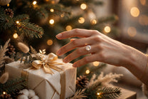 Woman's hand wearing a MARWAH lab-grown diamond engagement ring reaching for a Christmas gift beside a decorated Christmas tree