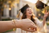 Joyful graduate throwing her cap in the air, wearing a gold MARWAH charm bracelet with graduation-themed charms