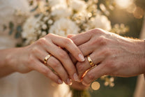 Newlywed couple's hands clasped together showing gold MARWAH wedding bands, with a white bridal bouquet glowing in soft sunlight